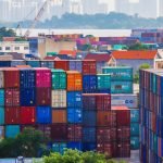 Vibrant stacked cargo containers at a bustling port with urban skyline in background.